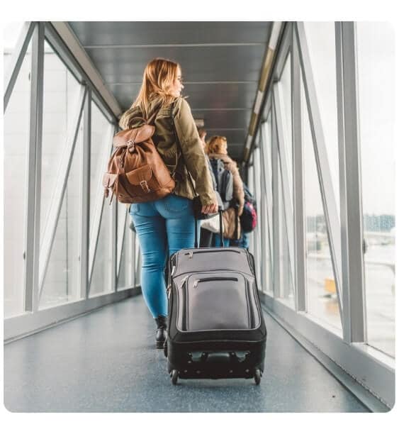 tanara blonda pe culoar in aeroport se indreapta spre imbarcare cu trolerul si priveste pe geam