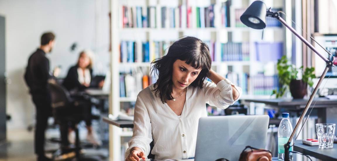 femeie tanara la birou ridica mana stanga la ceafa in timp ce lucreaza pe laptop si pe fundal doi colegi discuta