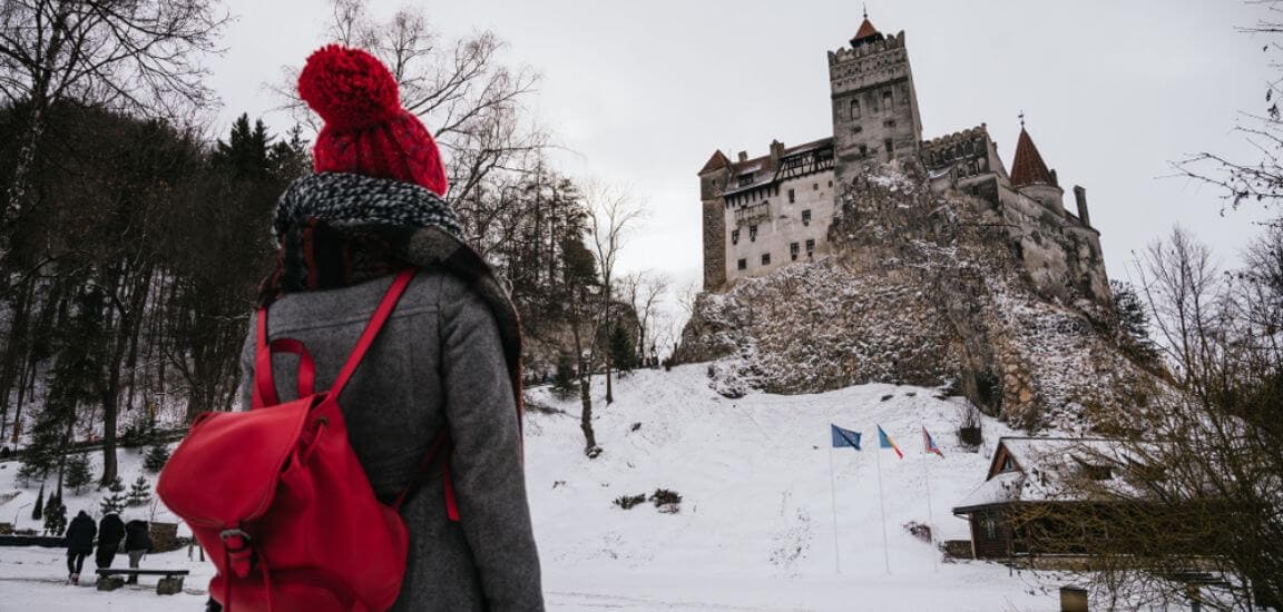 femeie tanara poarta o caciula rosie si priveste in departare castelul bran