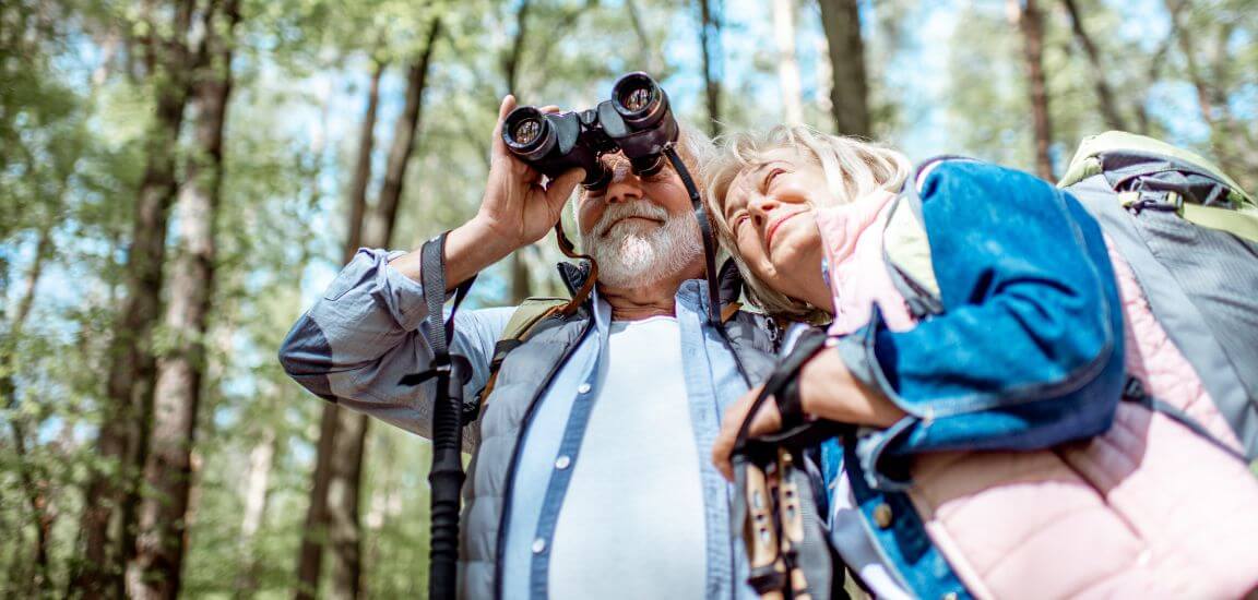 cuplu de pensionari se uita printr-un binoclu la pasari in padure