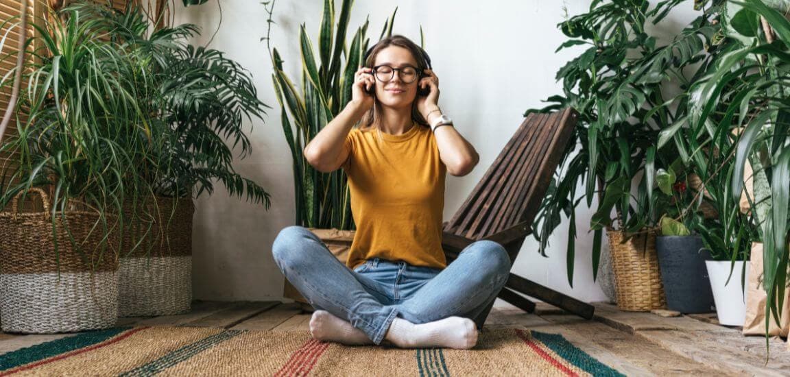 tanara intr-un tricou galben sta pe podea inconjurata de plante si asculta la casti un podcast de relaxare