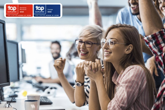 Un grup de colegi stau in birou in fata unui calculator si se bucura pentru reusitele lor